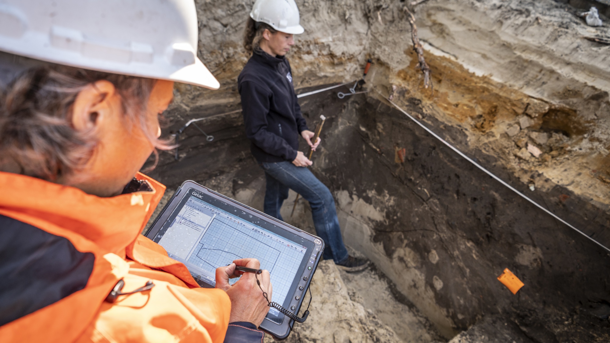 Mirjam staat in de kuil en Juliette staat er naast met een tablet in haar hand. Er wordt gekeken naar het zand.