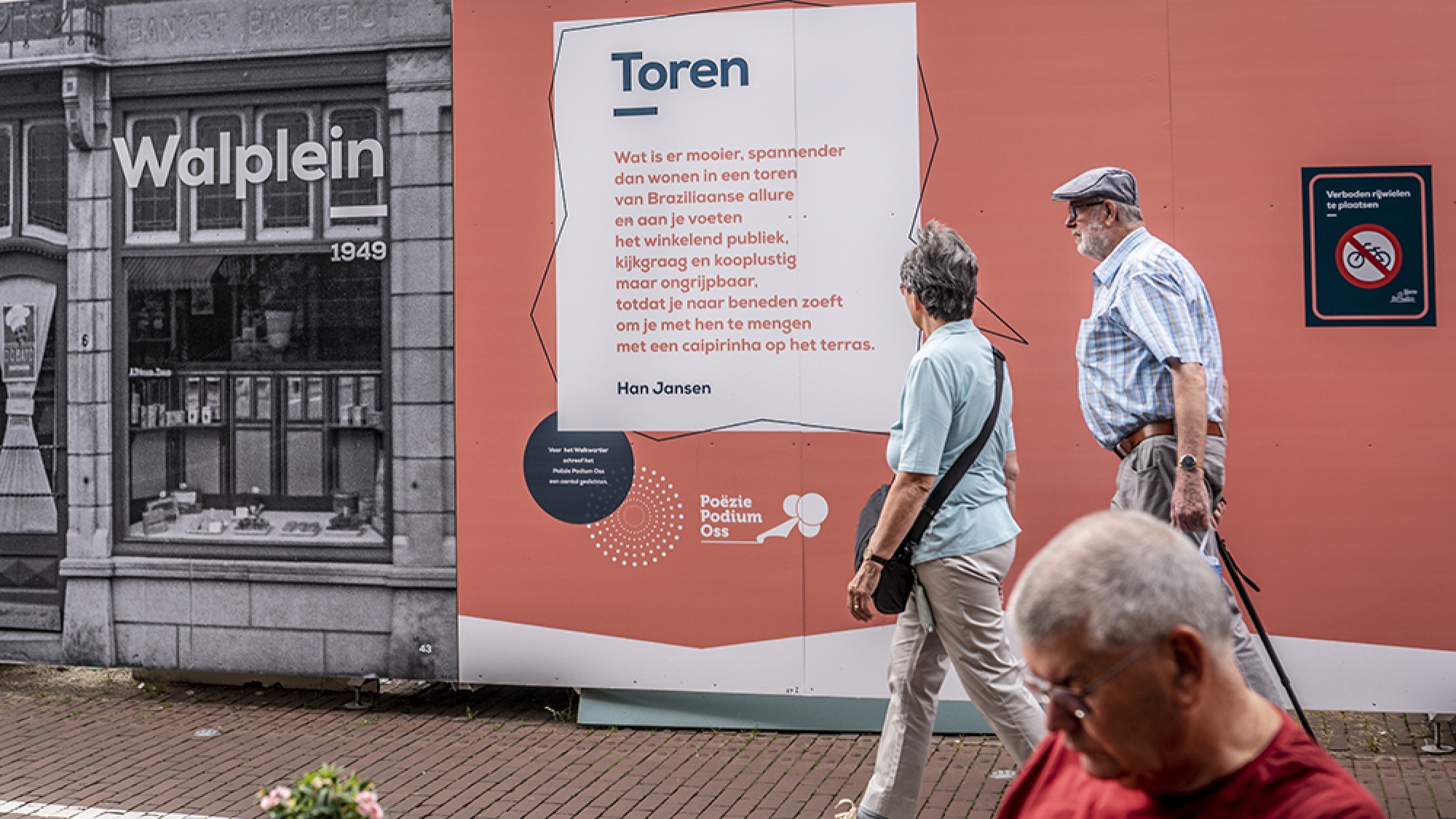 Een wat oudere man en vrouw lopen langs de bouwafrastering. Op de borden staat een tekst over de woontoren.