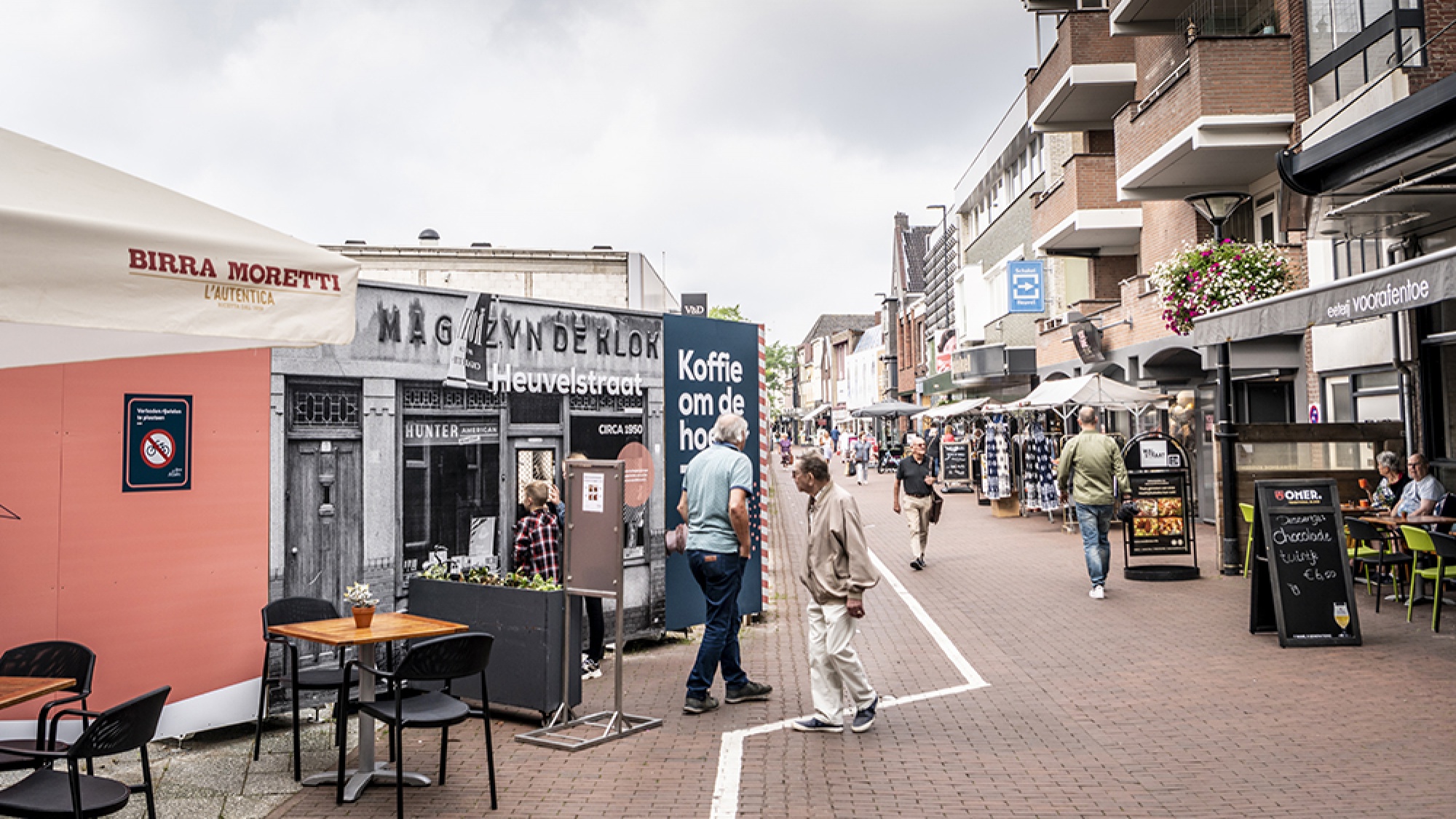 Foto van grotere afstand. Aan de linker zijde zie je de bouwafrastering en aan de rechter zijde restaurantjes. Hier tussenin lopen mensen rond.