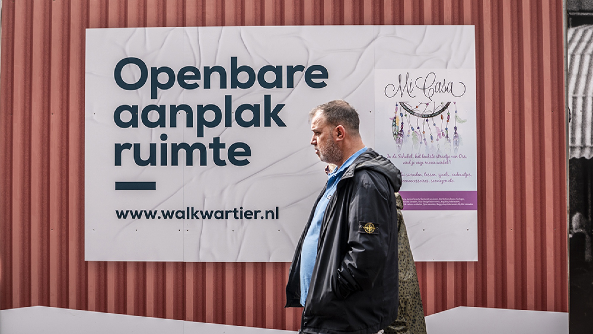 Een man loopt langs de bouwafrastering waar op staat: Openbare aanplak ruimte.