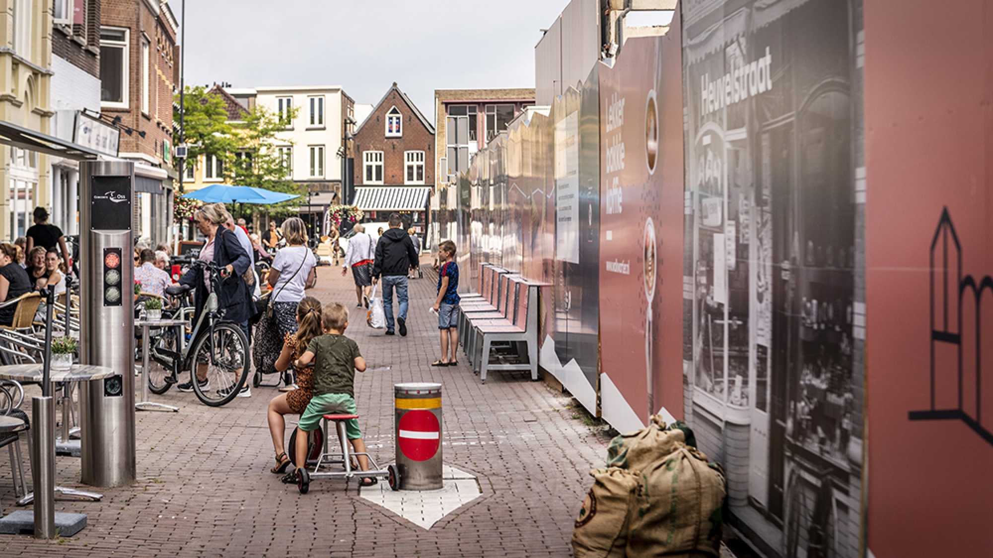 Aan de rechterzijde zie je de bouwafrastering en aan de linkerzijde zijn terrasjes. Hiertussenin lopen mensen. Ook zitten twee kinderen op een speelgoed auto.