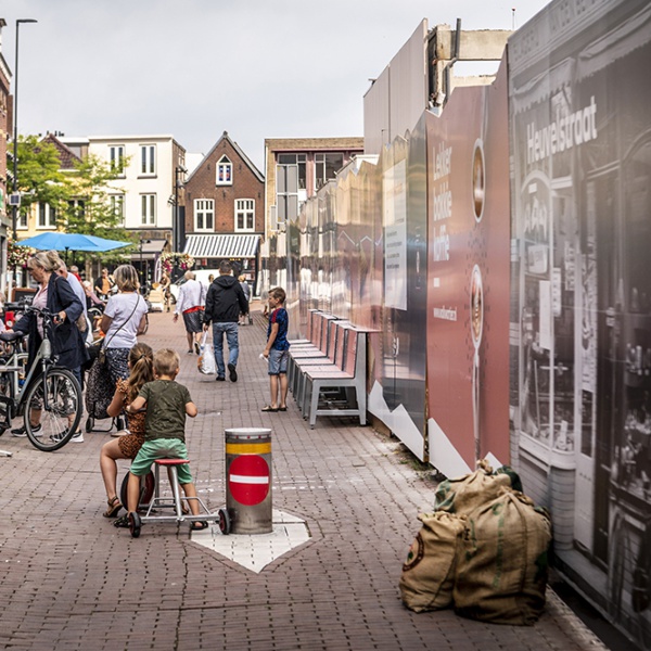 Een overzichtelijke foto waar aan de rechter zijde de bouwafrastering staat en aan de linker zijde volle terrassen zijn.