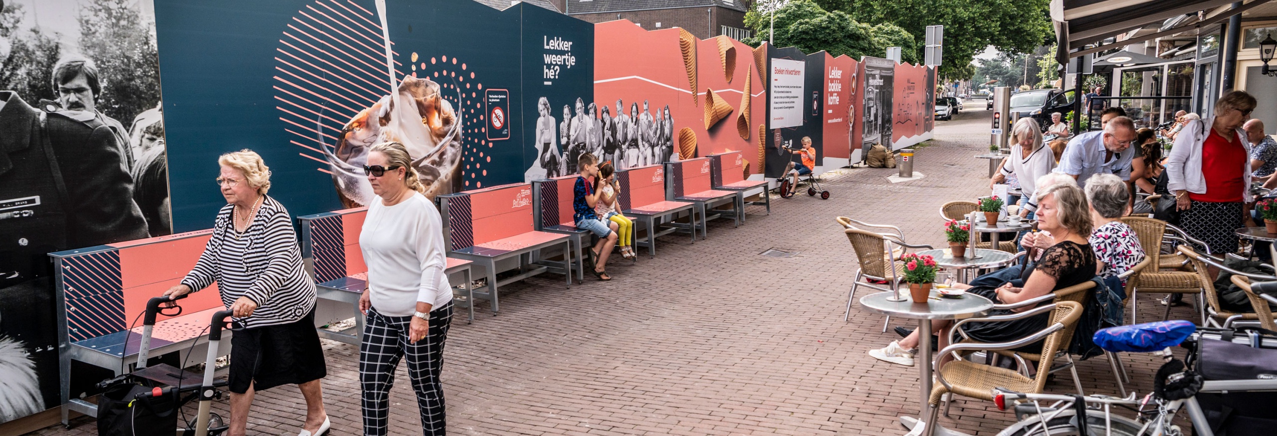 Aan de linkerzijde staat de bouwafrastering en aan de rechterzijde staan terrasjes. hier tussenin lopen mensen.